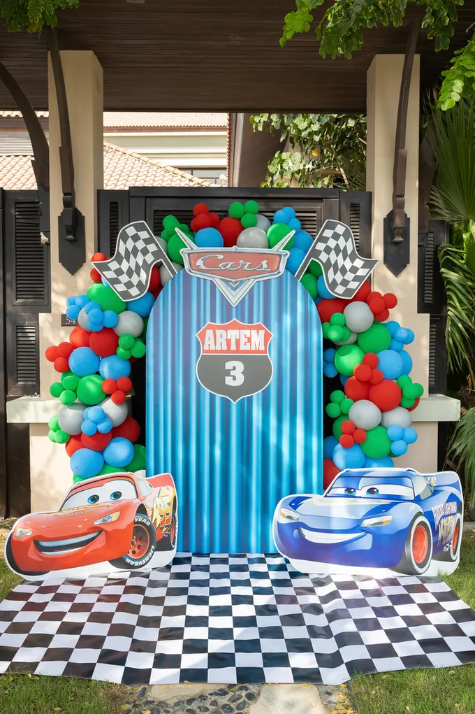 Cars-themed birthday backdrop featuring a blue striped panel that reads 'Artem 3', multicolored balloons, checkered floor, and life-size cutouts of cartoon cars (Lightning McQueen and another blue car).