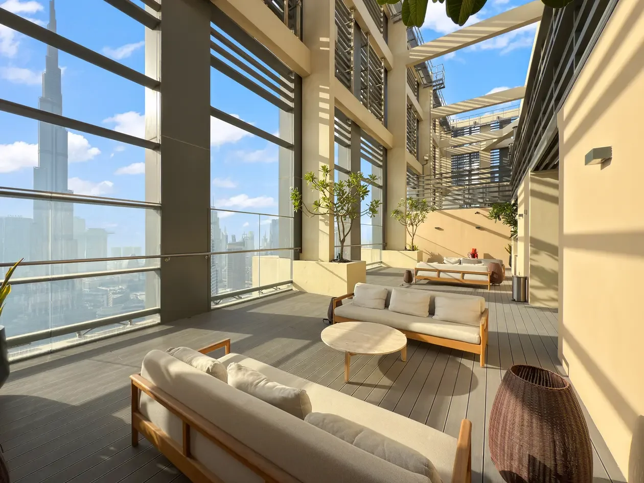 Rooftop lounge with beige cushioned sofas, round coffee tables, and potted trees overlooking a city skyline on a sunny day.