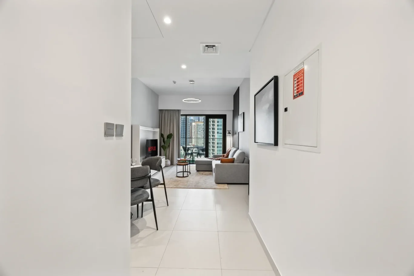Long hallway opening into a modern living room with white tiled floor, grey sofa, and floor-to-ceiling window view of city buildings.