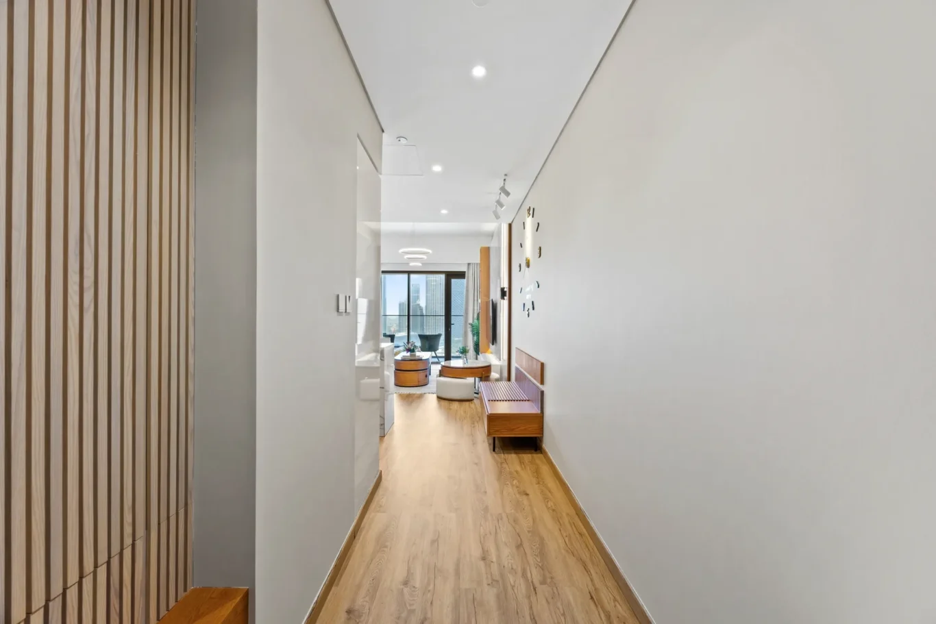 Long hallway with a wooden slat wall on the left, opening to a bright living area with round coffee tables and a city view through large windows.