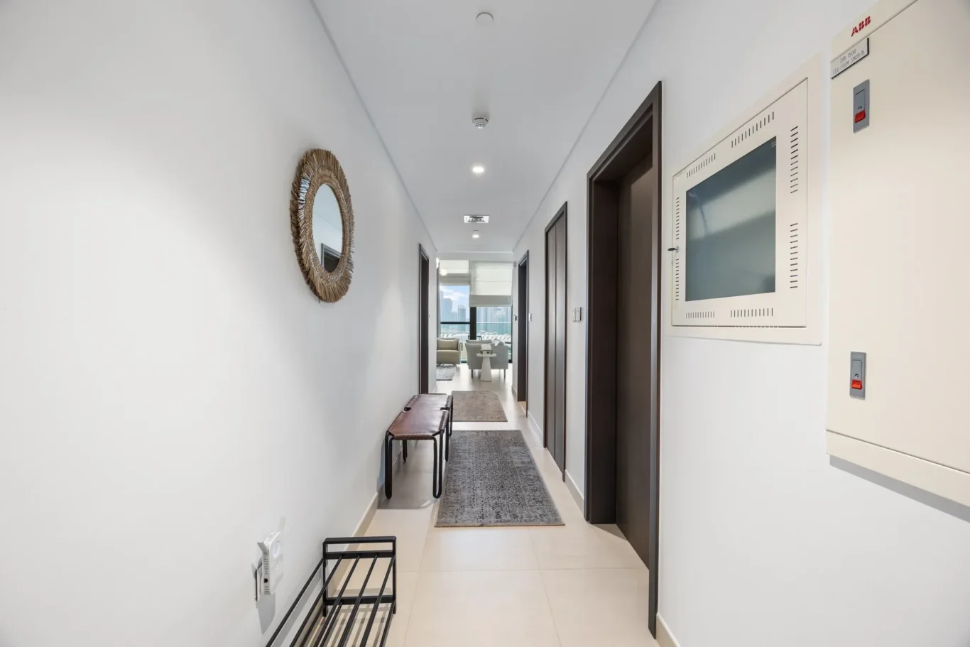 Bright, modern hallway with white walls, a round woven mirror on the left, a black metal bench, and gray runners; doors along the right and a living area visible at the end.