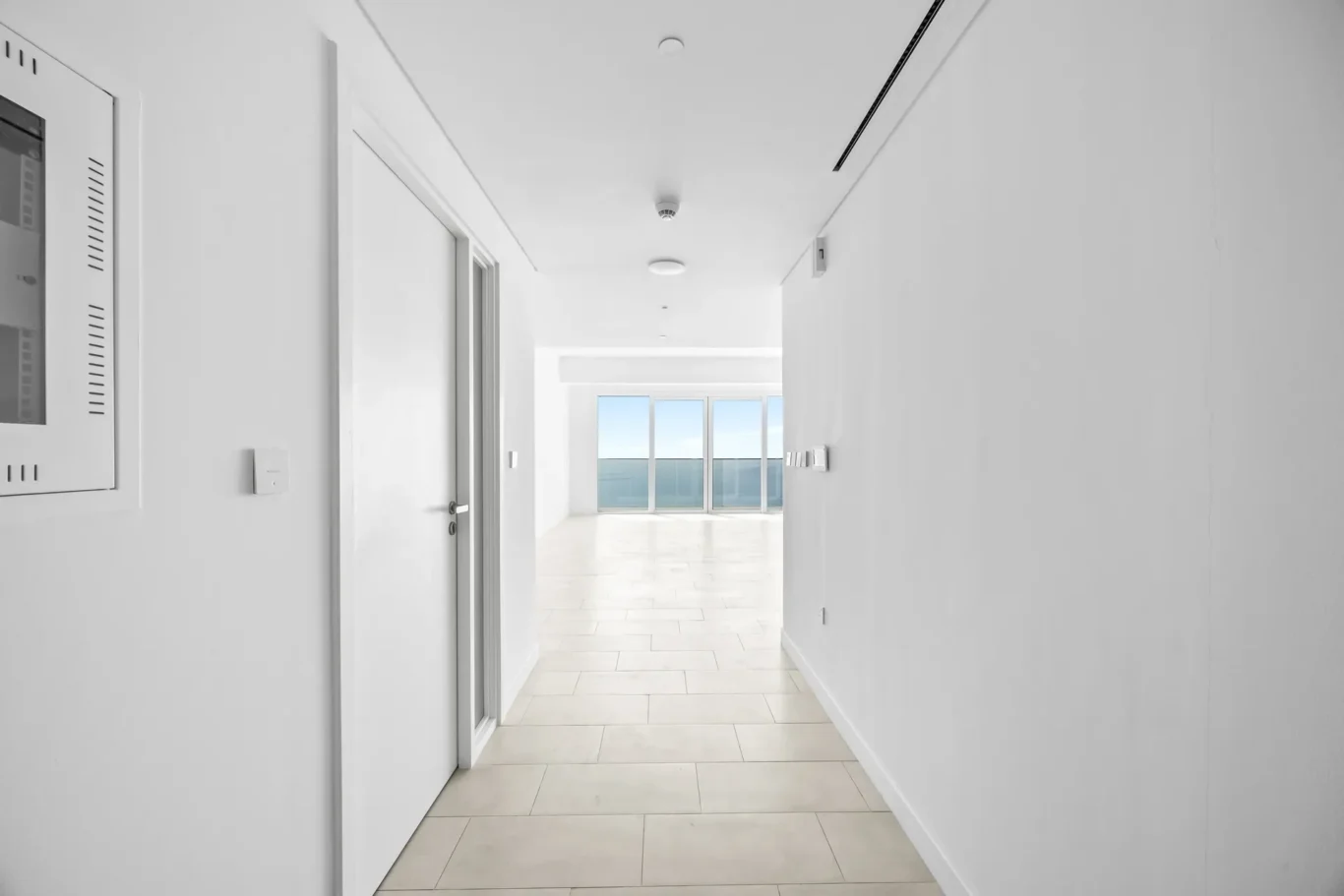 Bright white hallway with beige floor tiles leading to a sunlit balcony with large glass doors at the end.