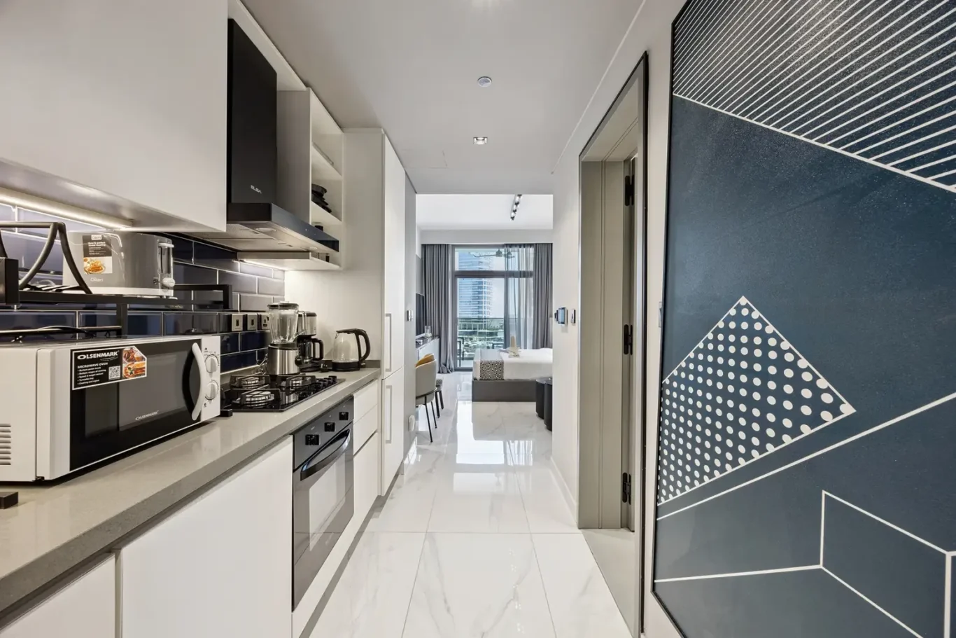 Modern galley kitchen with white cabinets, black backsplash, stainless appliances, and a marble floor leading to a living/sleeping area with a bed and balcony view.