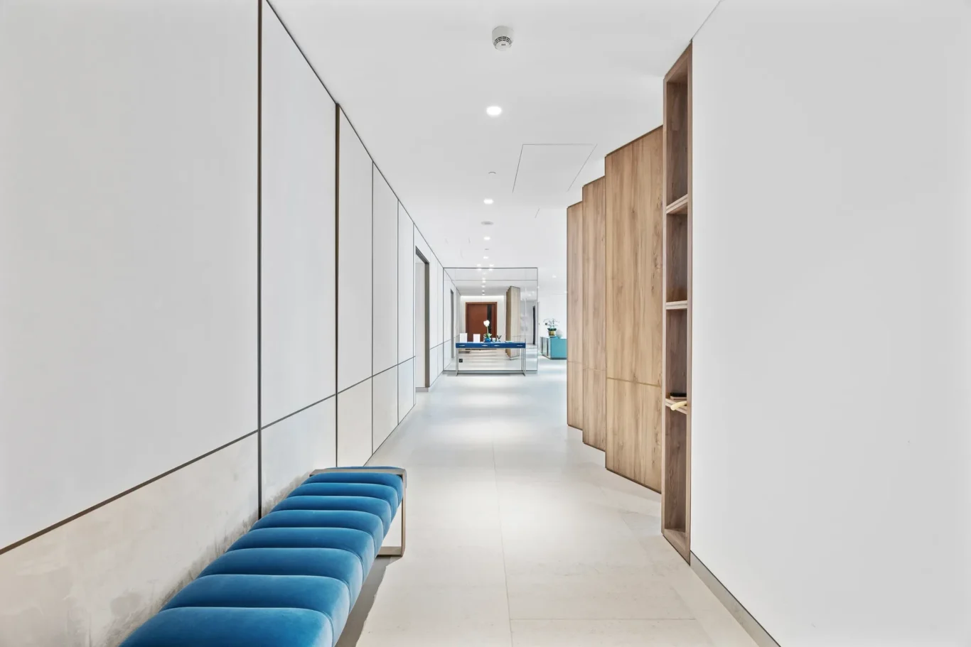Modern office hallway with white walls, long blue upholstered bench along the left, and wooden cabinets on the right.