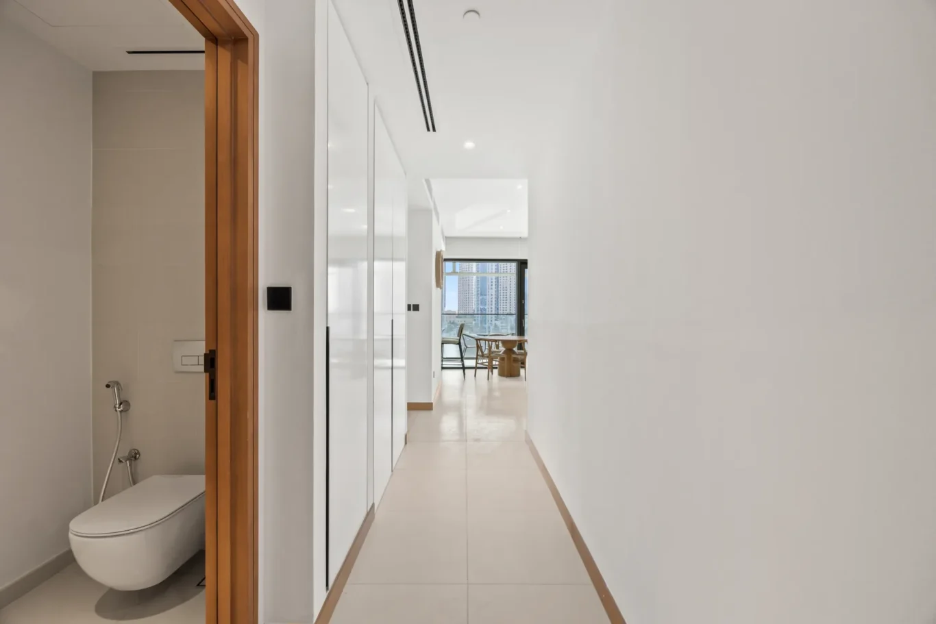 Bright modern hallway with white built-in cabinets on the left and a distant dining area with a city skyline through the window; bathroom with a wall-mounted toilet visible on the left.