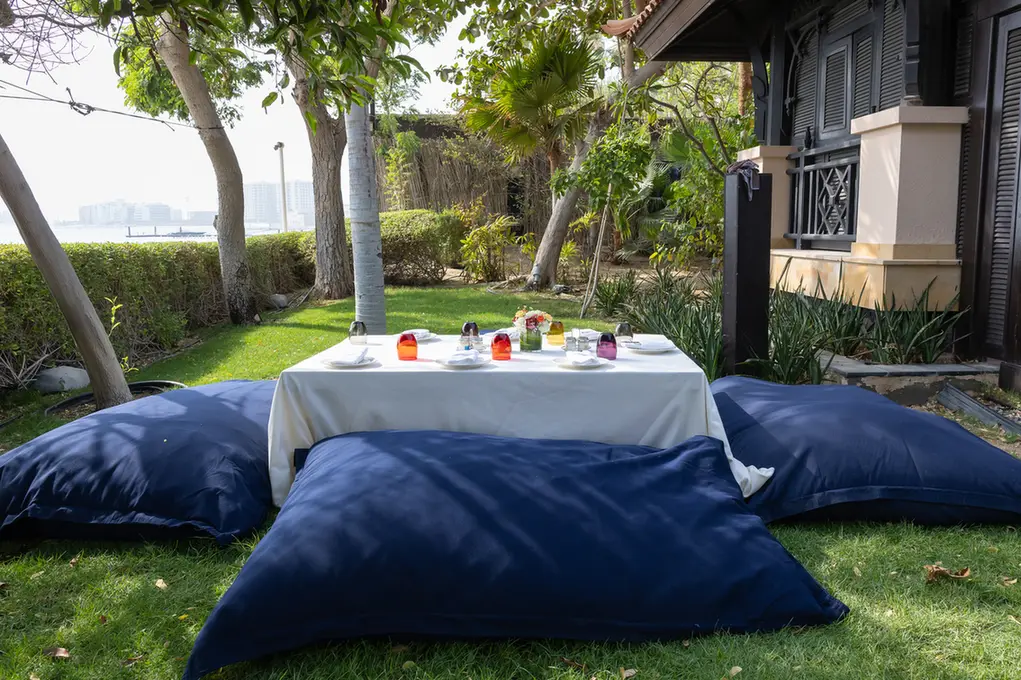 Outdoor garden dining setup with a low white table on a grass lawn, surrounded by large blue floor cushions.