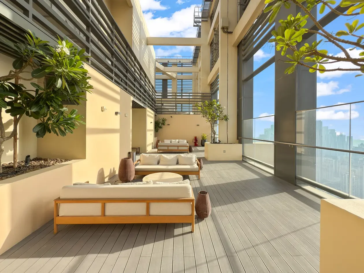 Rooftop lounge on a high-rise terrace with cream sofas and wooden frames, potted trees, and a glass railing overlooking a city skyline.