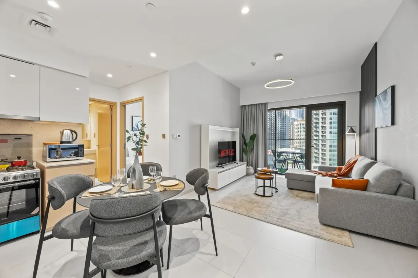 Open-concept living area with a white kitchen on the left, round dining table, gray chairs, and a seating area facing a TV showing Netflix. A balcony and city skyline are visible through sliding doors.