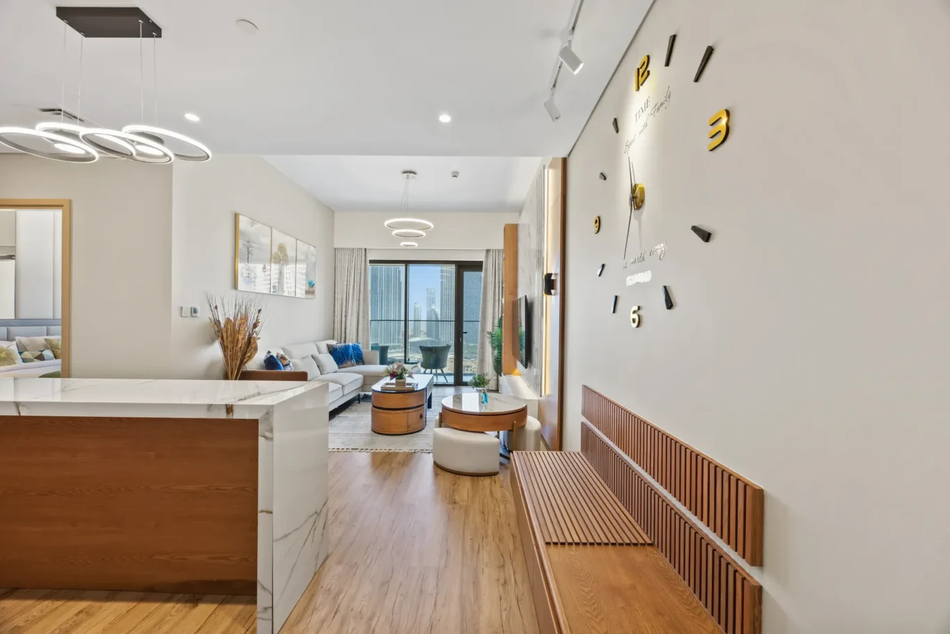 Modern living room with a large wall clock, white sofa, and city skyline visible through glass doors on the far end.
