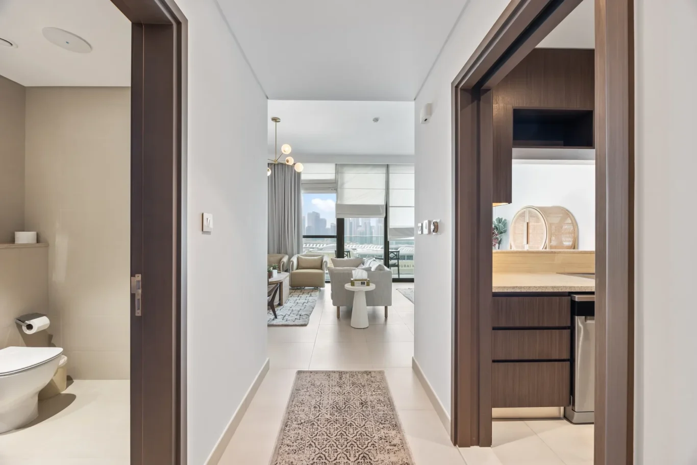 Open hallway leading to a bright living room with city skyline visible through large windows at the end. Living room at the end of a hallway with grey seating and floor-to-ceiling windows overlooking the city. Modern apartment entry hall with a patterned rug, white walls, and dark wood cabinetry on the right, opening to a sunny living area. Bright, contemporary interior: hallway to living room with a city view and stylish furniture.