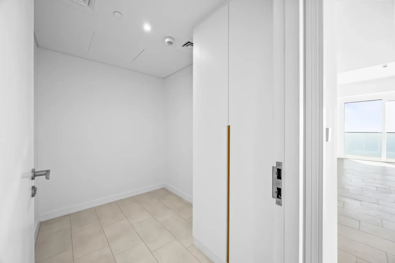 Bright white hallway with beige floor tiles and a doorway revealing a sunlit room with a balcony and view of the sea/ocean.