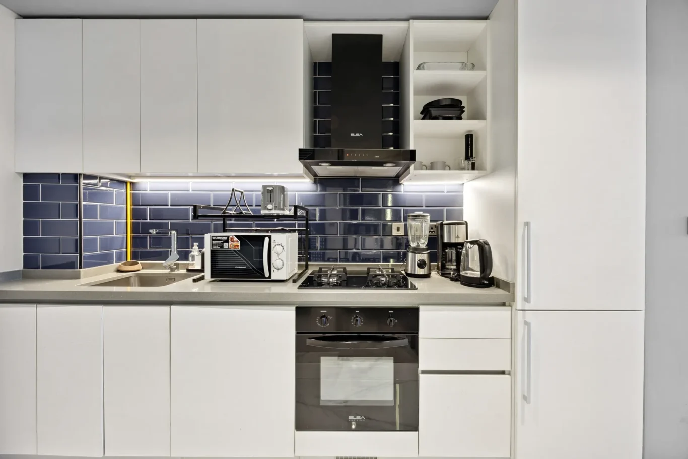 White kitchen with dark blue tile backsplash, built-in oven, and open shelves holding bowls and glasses.