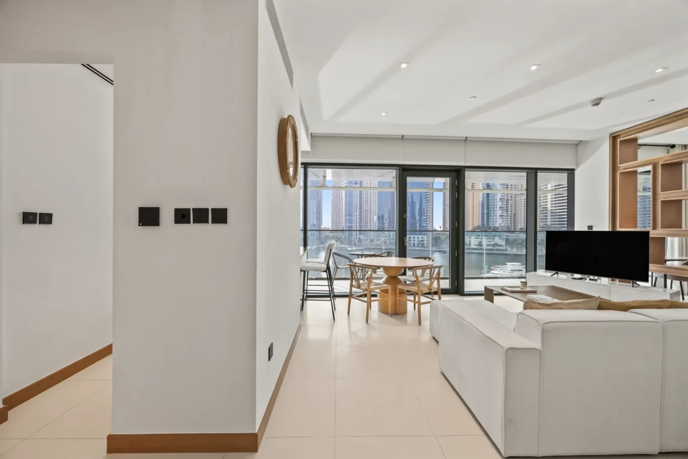 Bright, open-plan living area with a white sectional sofa, wooden dining table, and large glass doors overlooking a city skyline.