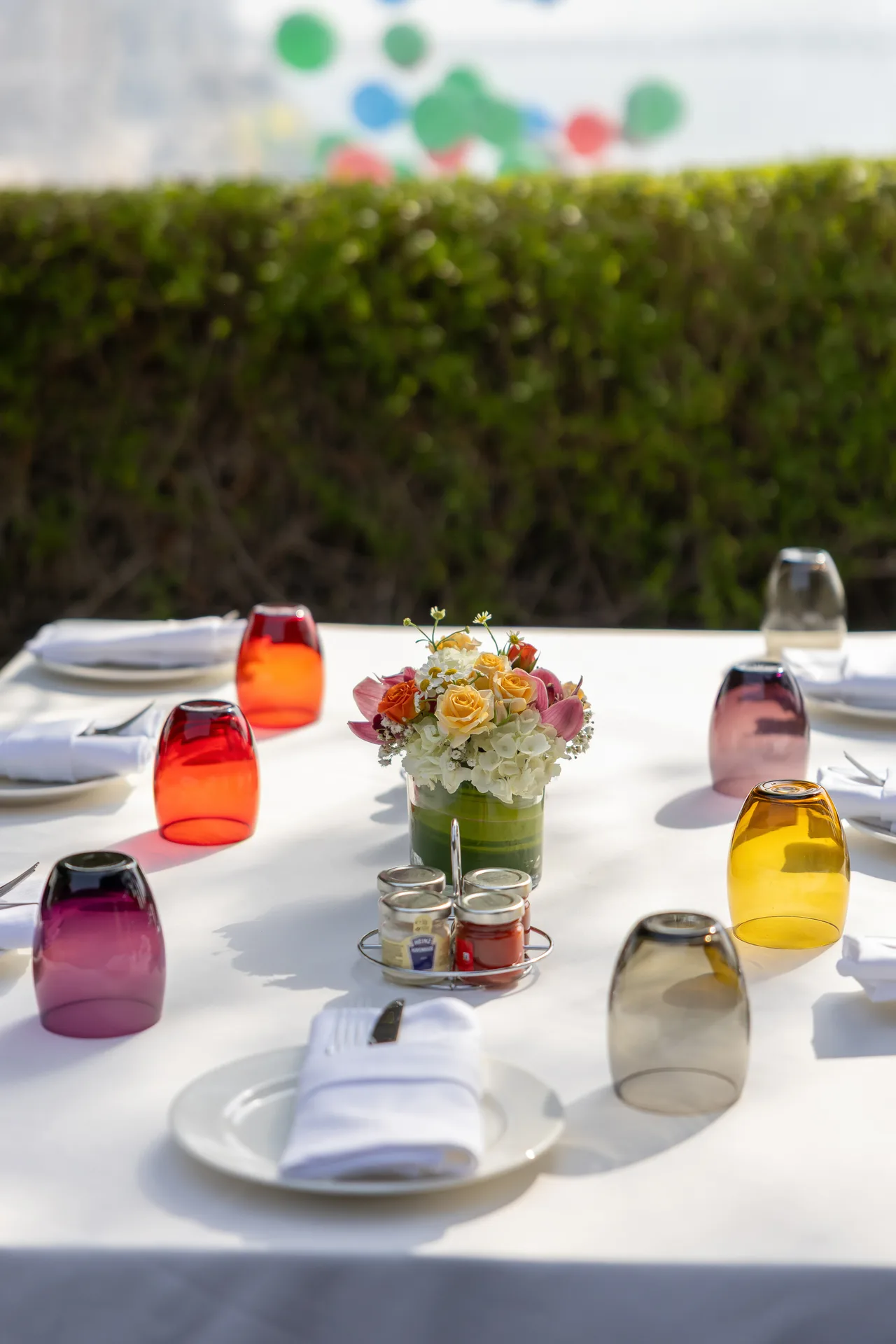 Outdoor dining table with a white tablecloth, colorful glassware, and a bright floral centerpiece; green hedge and blurred balloons in the background.