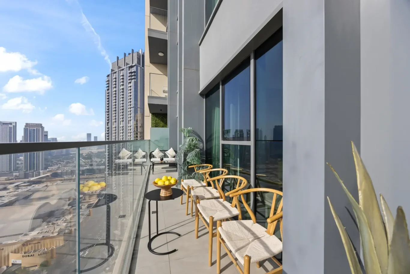 Balcony with wooden chairs and a small table holding lemons, glass railing, overlooking a city skyline of tall buildings.