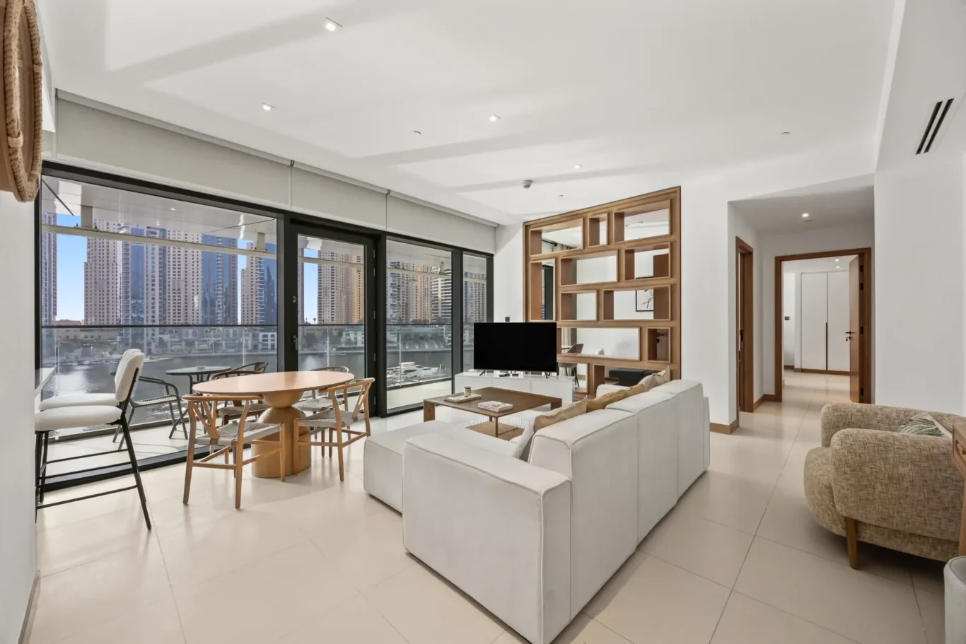 Bright open-plan living room with white sofas, wooden shelving divider, and city skyline visible through large glass doors to the balcony.