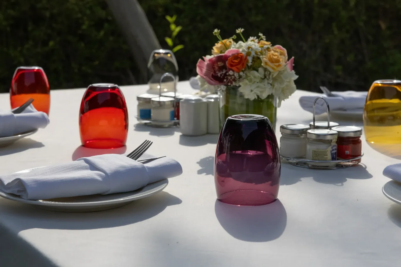 Outdoor dining table set with white plates, folded napkins, and colorful glass goblets; floral centerpiece.