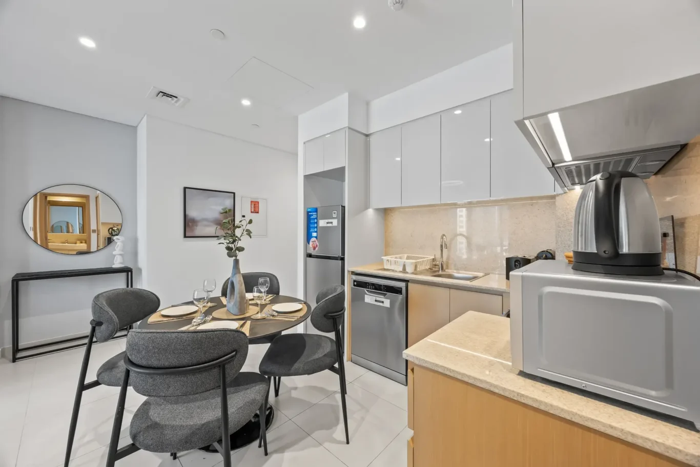 Open-plan kitchen and dining area with white glossy cabinets, stainless steel appliances, and a round dining table set for four with gray chairs.