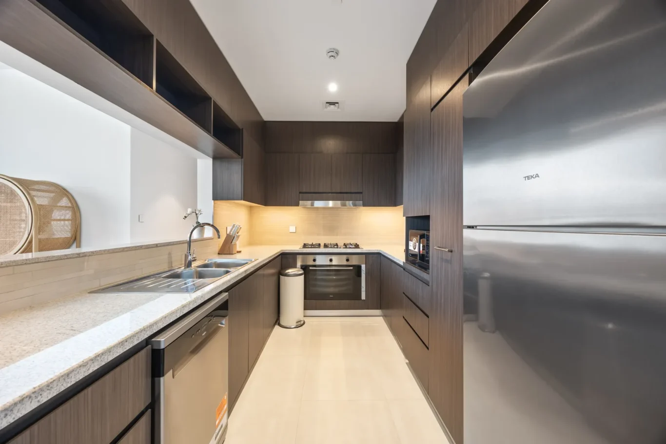 Narrow modern galley kitchen with dark wood cabinets, beige countertops, and stainless steel appliances.",