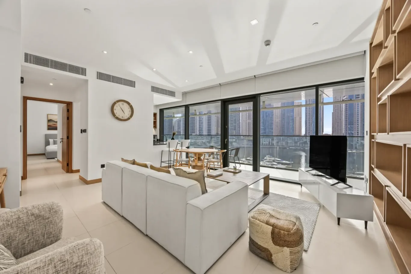 Bright, modern living room with a sectional sofa, woven ottoman, and a large glass wall overlooking a marina and city skyline nearby.