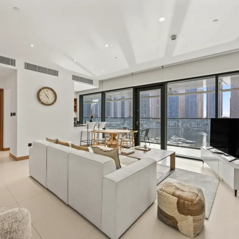 Bright, modern living room with a sectional sofa, woven ottoman, and a large glass wall overlooking a marina and city skyline nearby.