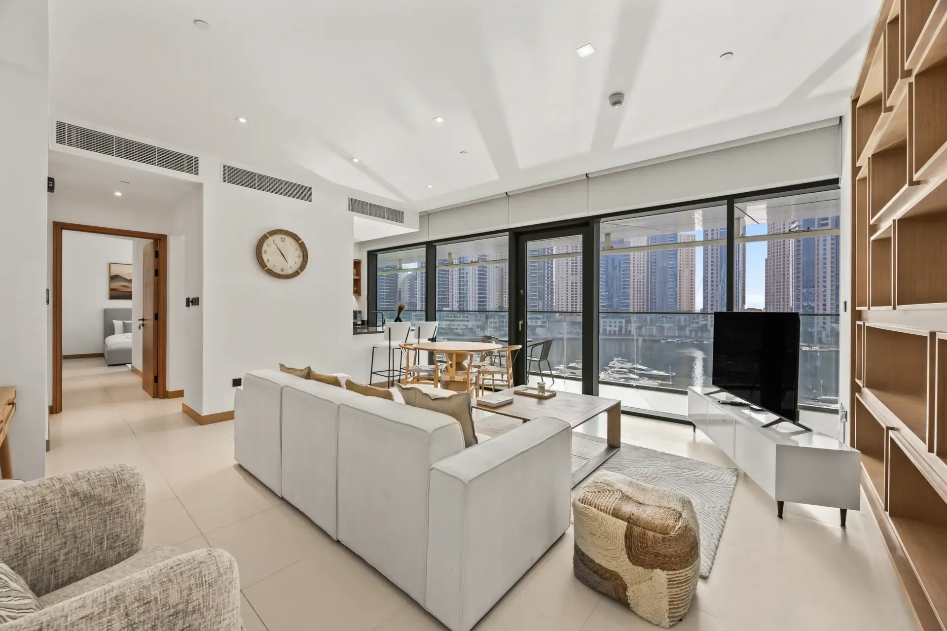 Bright, modern living room with a sectional sofa, woven ottoman, and a large glass wall overlooking a marina and city skyline nearby.