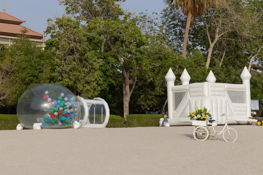 A transparent inflatable bubble with multicolored balls next to a white inflatable tunnel in a park setting.