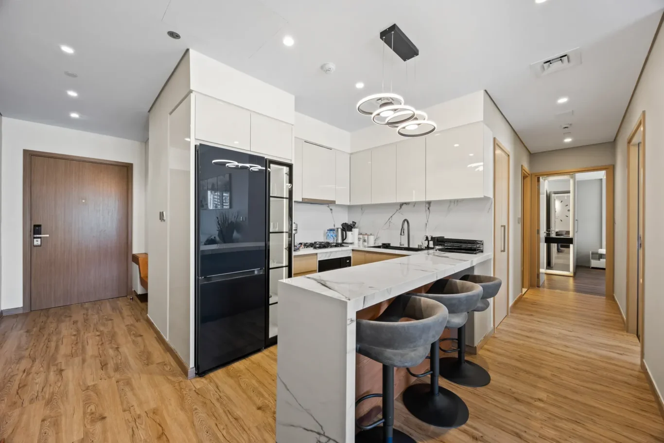 Modern kitchen with white gloss cabinets, marble island, and a black refrigerator; four gray bar stools.