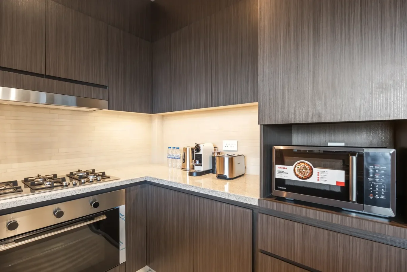 Modern L-shaped kitchen with dark wood cabinets, beige backsplash, and stainless appliances including stove, oven, microwave, and toaster.