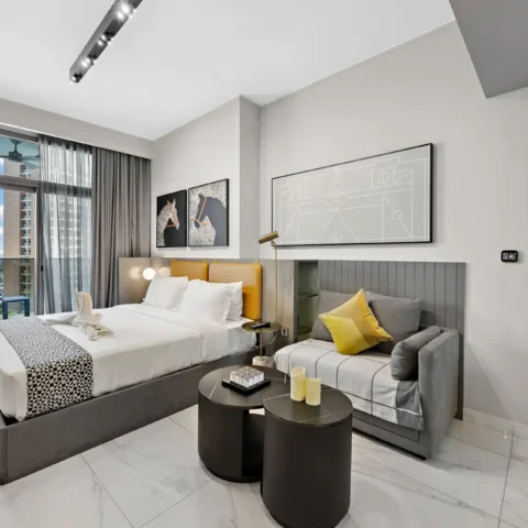 Modern hotel room with a large bed, grey headboard, yellow accent pillows, and a grey sofa near artwork on the wall.
