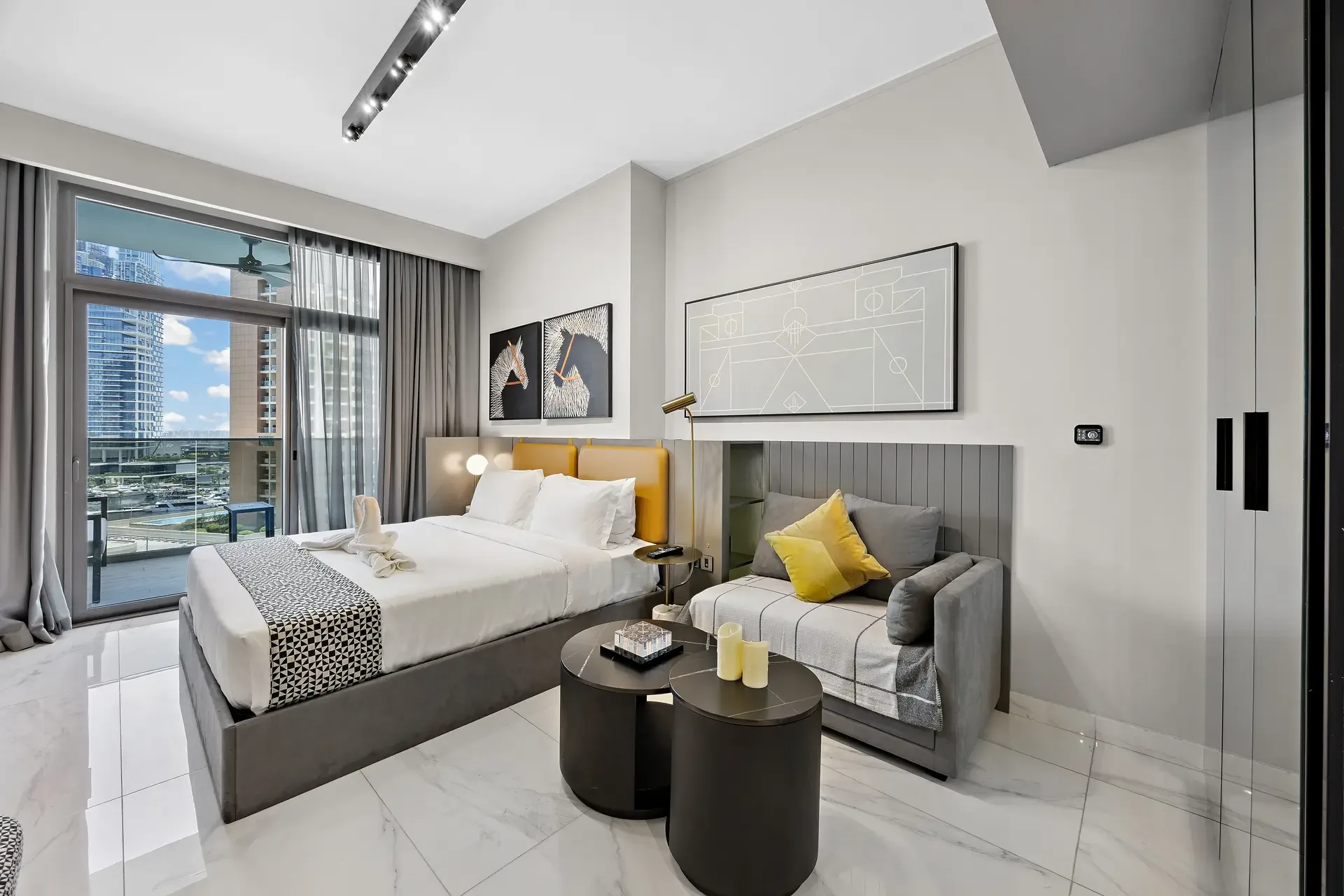 Modern hotel room with a large bed, grey headboard, yellow accent pillows, and a grey sofa near artwork on the wall.