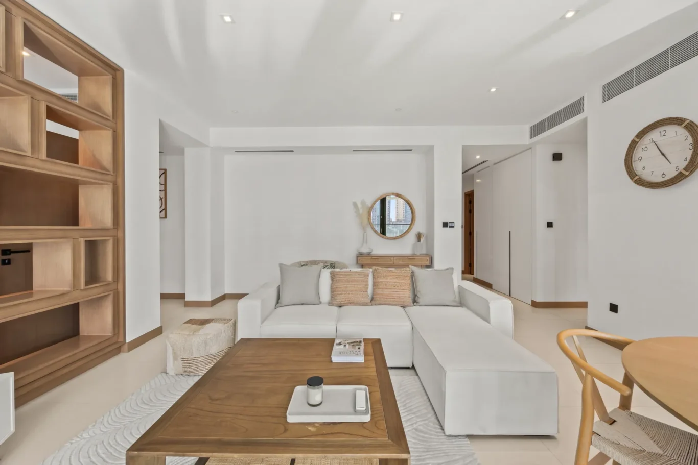 Bright open living room with white sectional sofa, wooden coffee table, and beige pillows; large round mirror and console table against white walls.