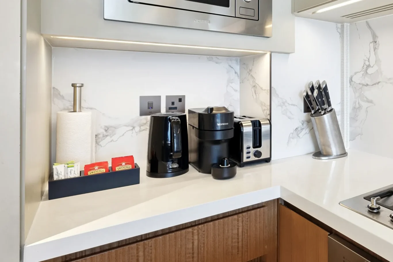 Modern kitchen counter with a coffee maker, Nespresso machine, toaster, knife block, and tea sachets against a marble backsplash.