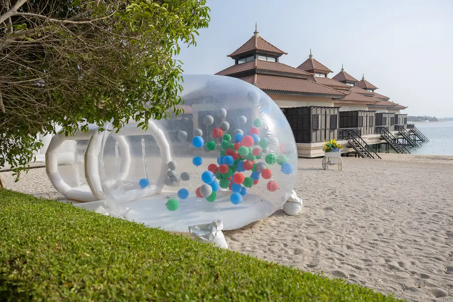 Transparent inflatable bubble filled with colorful balls on a sandy beach, with overwater cottages in the background.