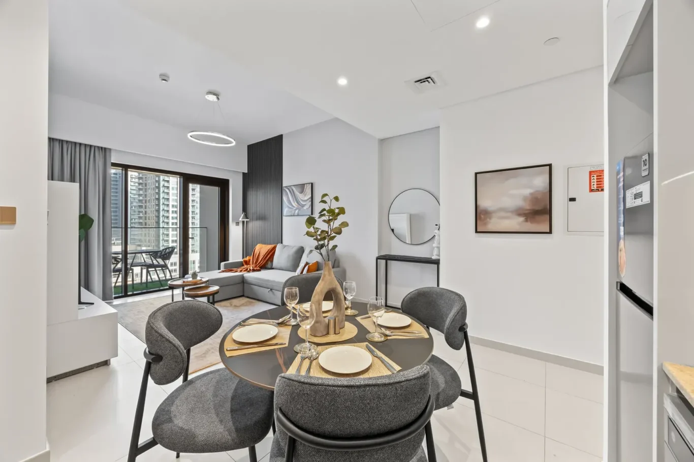 Open-plan living and dining area with a round dining table set for four, gray chairs, and a sofa by a balcony overlooking city buildings.