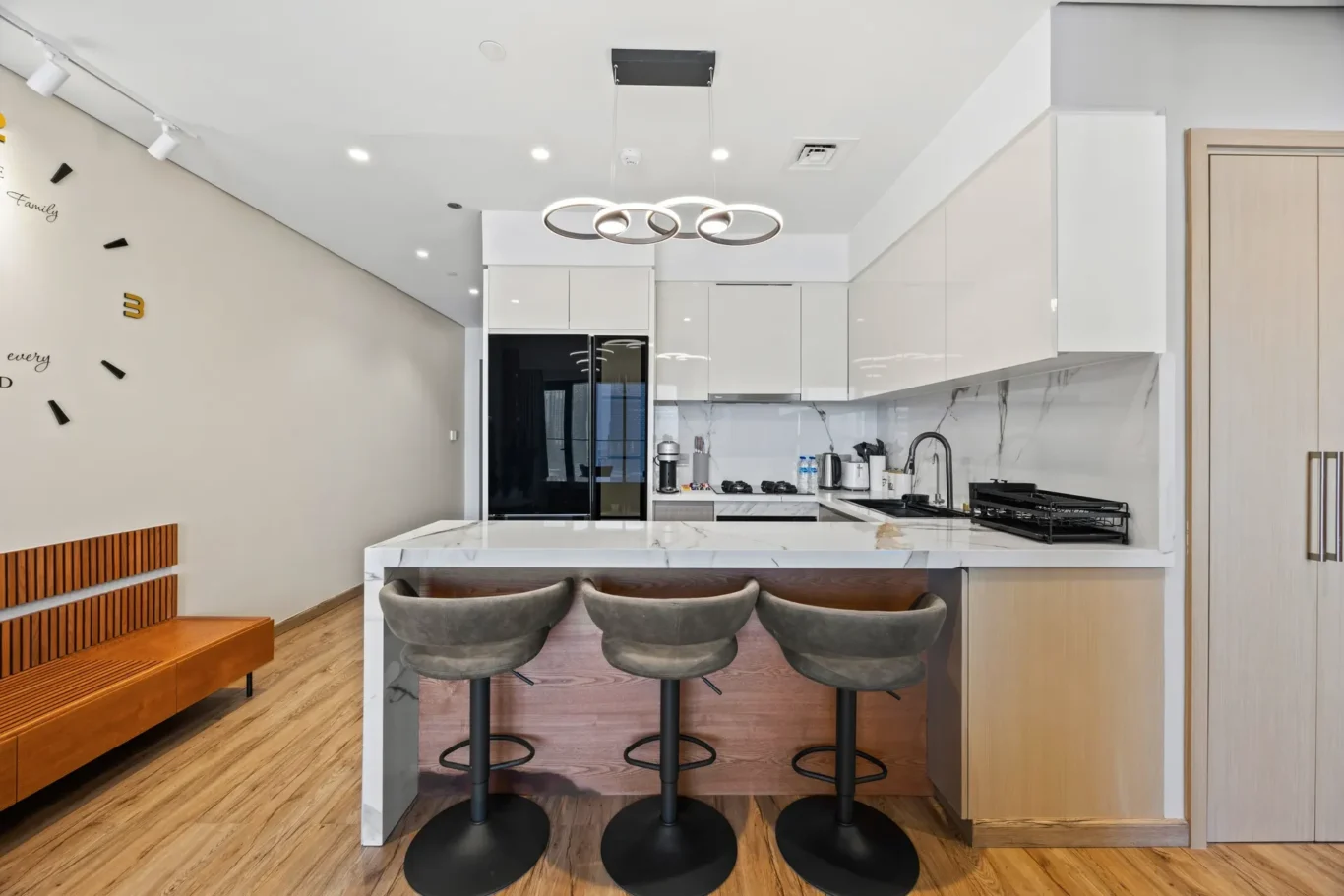 Modern white kitchen with marble-topped island, three black pedestal stools, and a circular pendant light above.