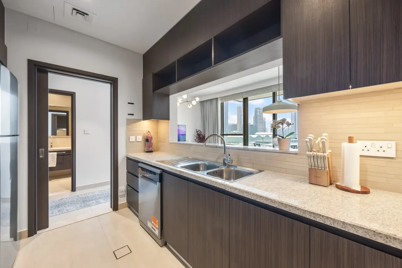 Modern kitchen with dark wood cabinets, double sink, and stainless dishwasher along a speckled countertop; open pass-through to living area with city view outside the window.