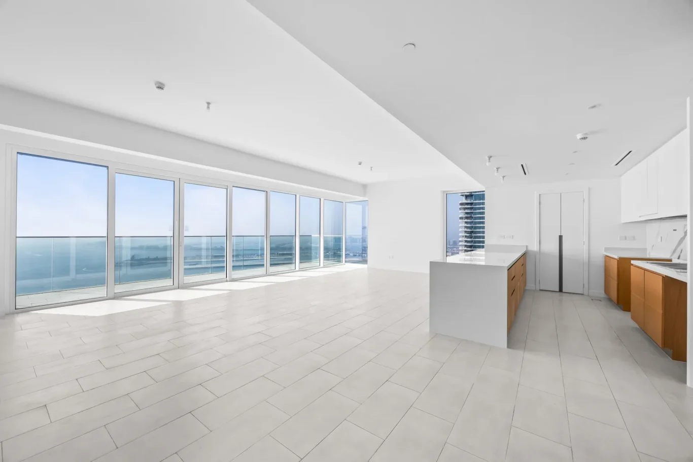 Open-plan kitchen and living area with floor-to-ceiling glass doors opening to an ocean-view balcony; white surfaces and light tiles.