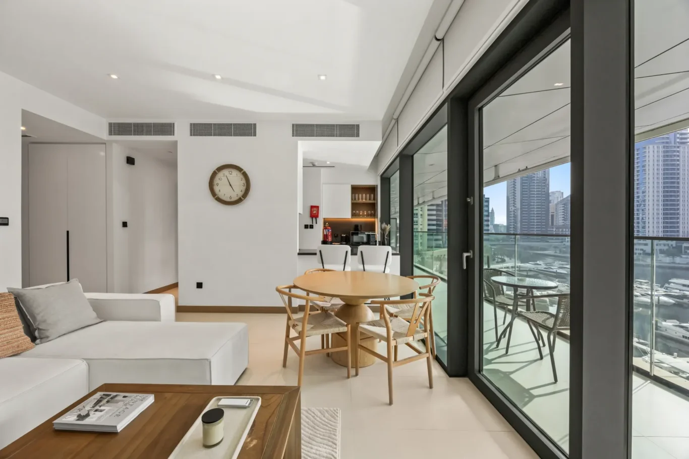 Bright open-plan living area with a white sofa, wooden coffee table, round dining table, and floor-to-ceiling glass doors opening to a balcony with a city skyline view.