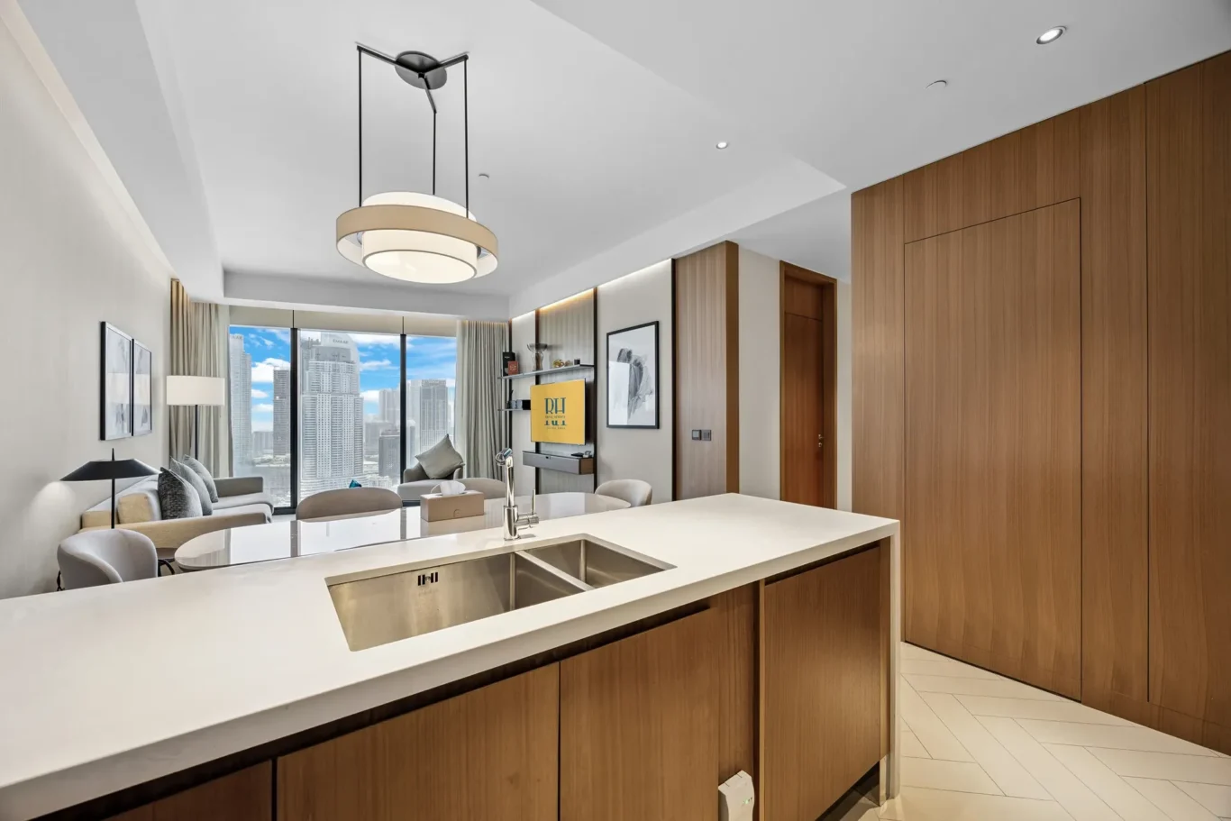 Open-concept kitchen and living area in a modern apartment with wood cabinets, white countertops, and a city skyline visible through large windows.