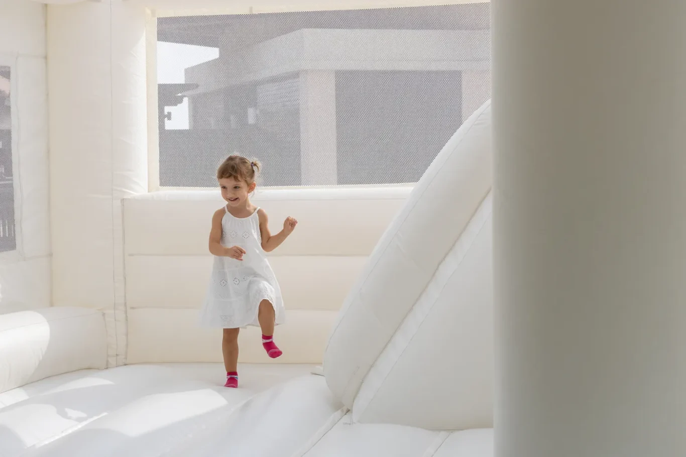 A young girl in a white dress jumps and smiles inside a bright white inflatable bounce house.