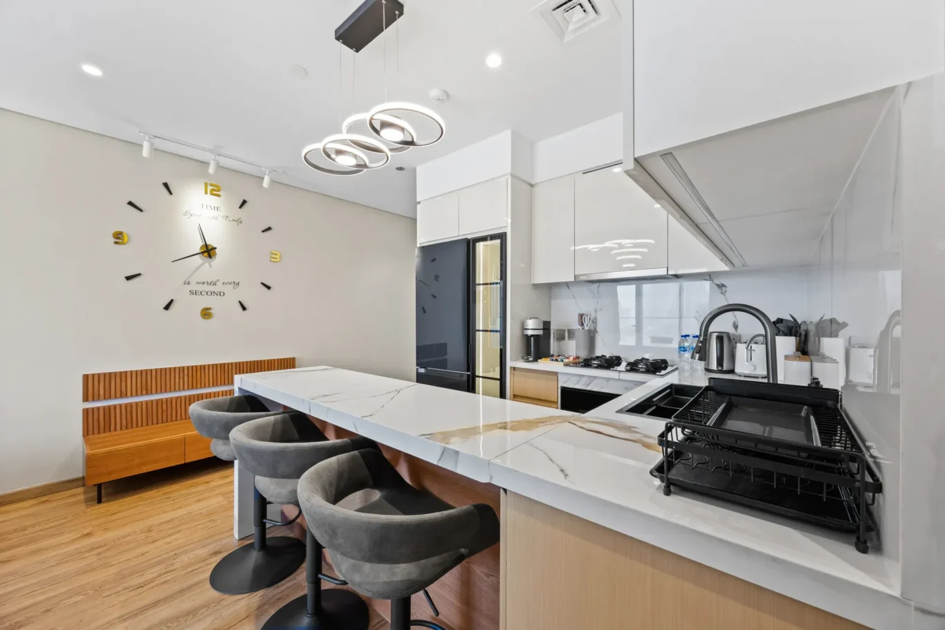 Modern kitchen with marble island, four gray swivel stools, white cabinetry, and a large decorative wall clock with gold numerals on the wall above a wooden bench.