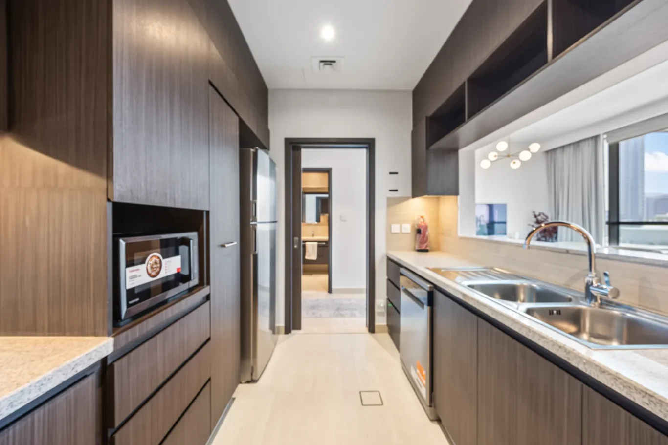 Modern galley kitchen with dark wood cabinets, stainless appliances, and a double sink along an island facing a window view of the city.