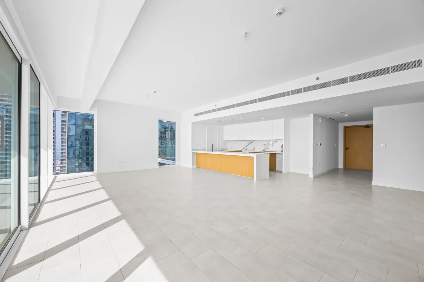 Bright open-concept living area with white walls, large glass doors, and a yellow kitchen island overlooking the city from high windows