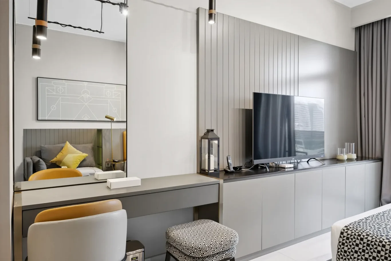 Modern hotel room scene with a grey panel wall, a mounted flat-screen TV, and a desk reflecting a bed with yellow and grey cushions in a mirror.