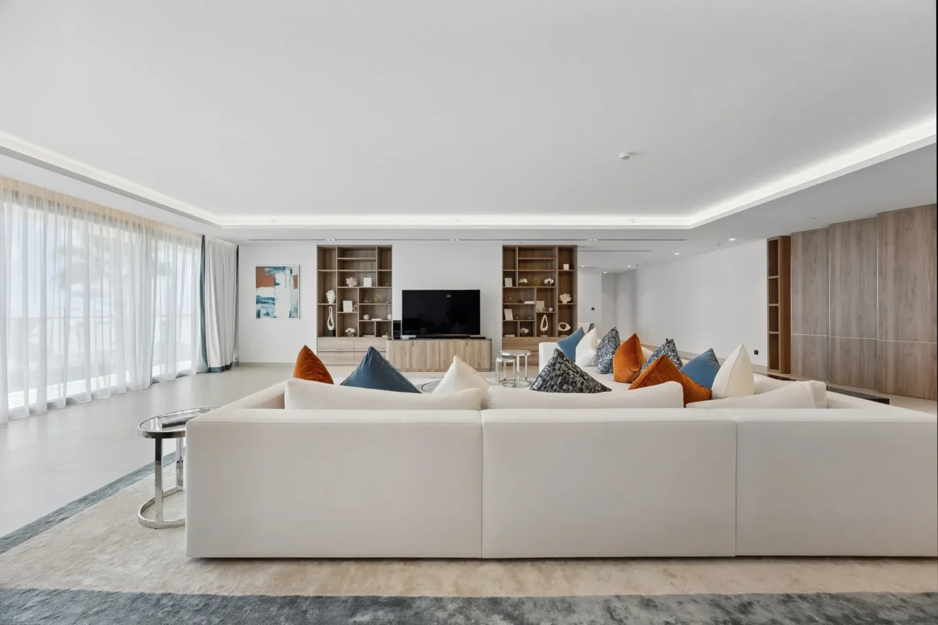 Bright modern living room with a white sectional sofa facing a TV on wood shelving, colorful throw pillows, and sheer-curtained floor-to-ceiling windows on the left.
