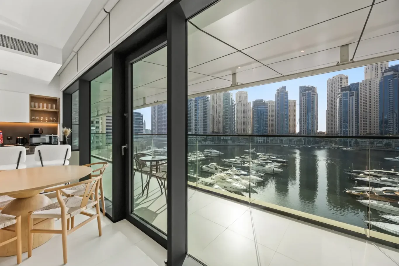 Open-plan kitchen and dining area with glass doors opening to a balcony overlooking a marina and city skyline with high-rise buildings and yachts.