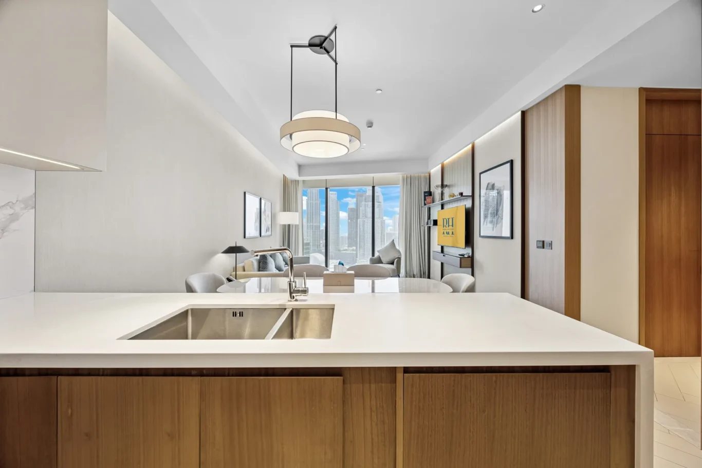 Modern kitchen island with a built-in sink in foreground and warm wood cabinetry below; bright, open living area beyond with city skyline through large windows.