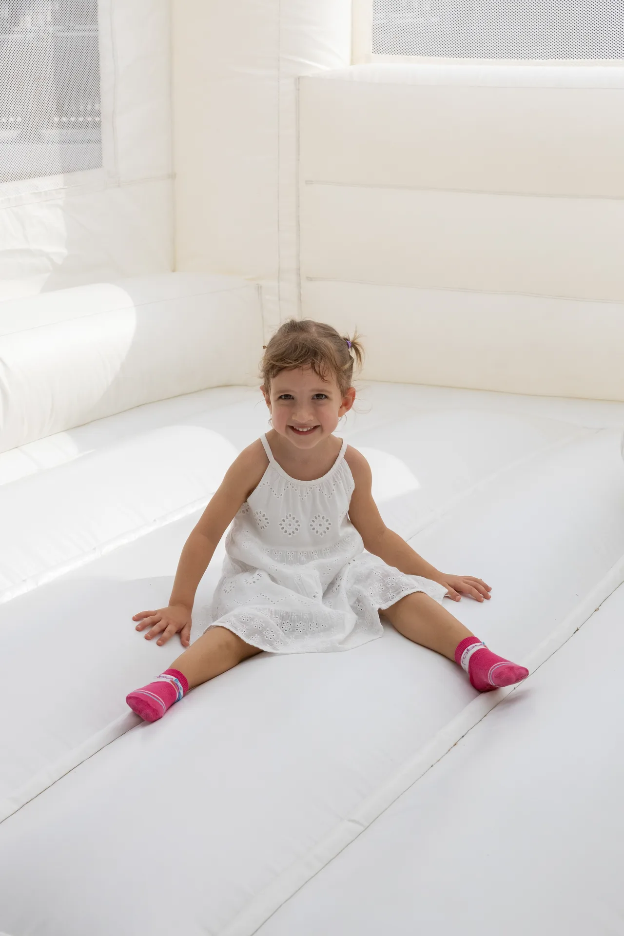 Smiling young girl in a white dress sitting on a white inflatable slide/sofa surface and looking at the camera with pink socks showing on both feet.