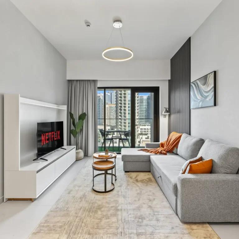 Modern living room with a gray sectional sofa, orange cushions, and a white media console hosting a Netflix TV. A balcony with city views is visible outside.